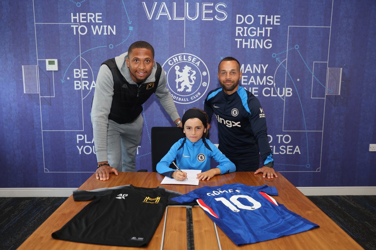 Young player signing at Chelsea between two coaches, a Monster Mentality top and a Chelsea shirt on the table