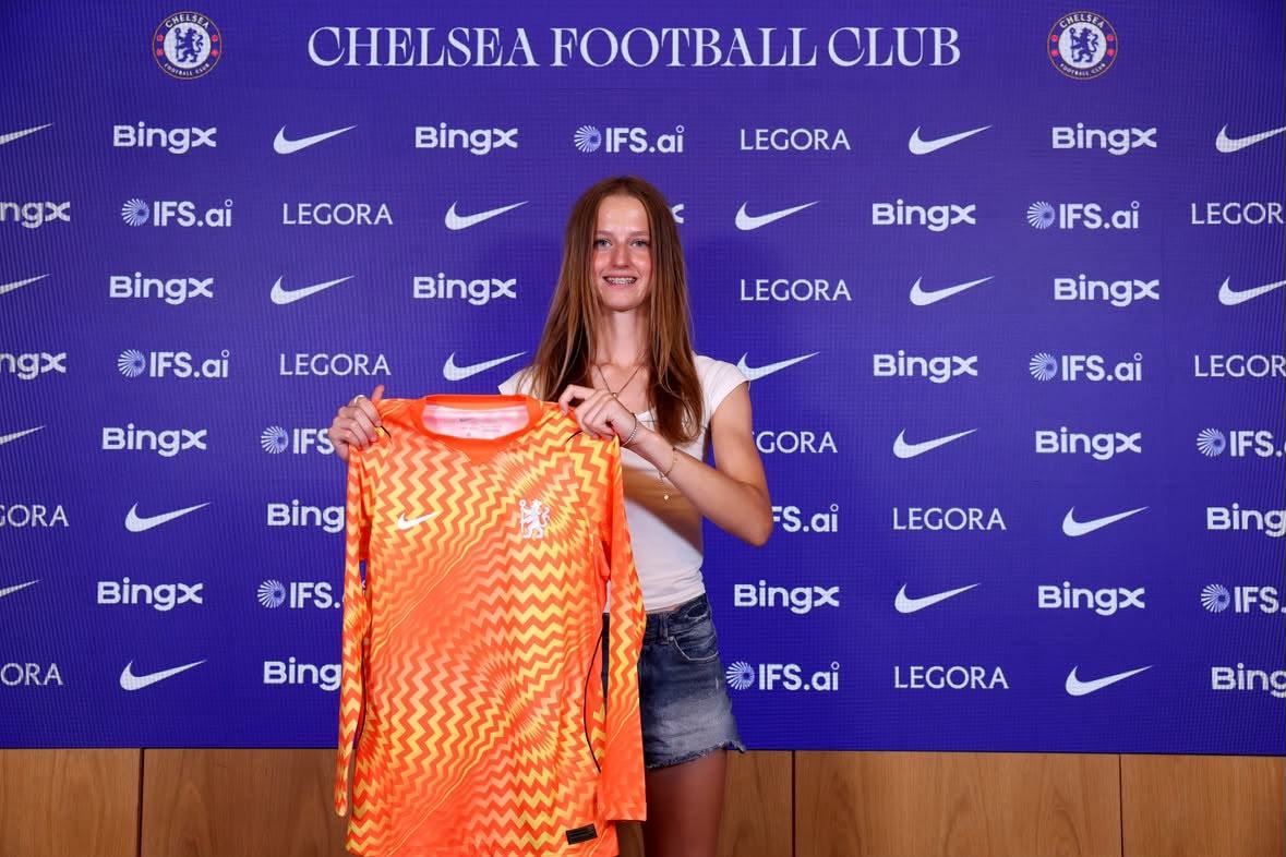 Young player holding up an orange Chelsea goalkeeper shirt in front of the Chelsea Football Club backdrop