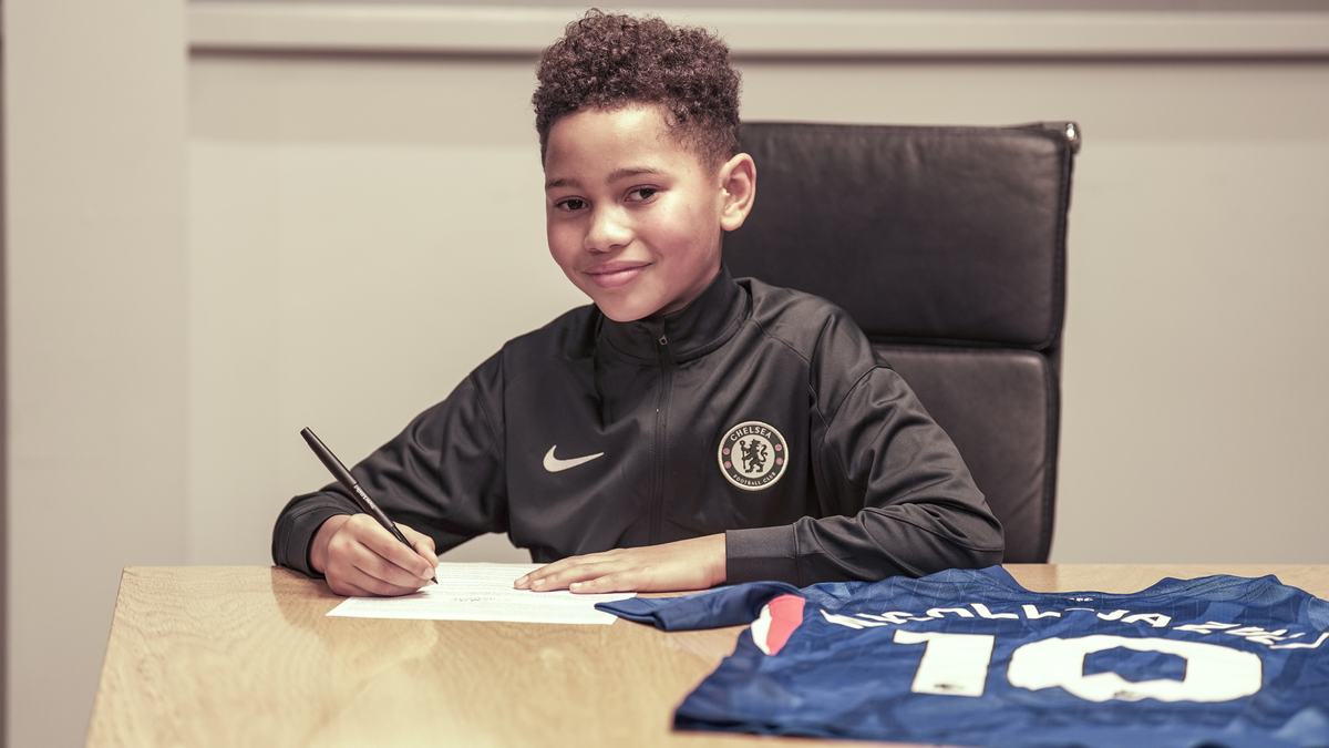 Smiling young player in a Chelsea tracksuit signing at a desk, a Chelsea shirt beside him
