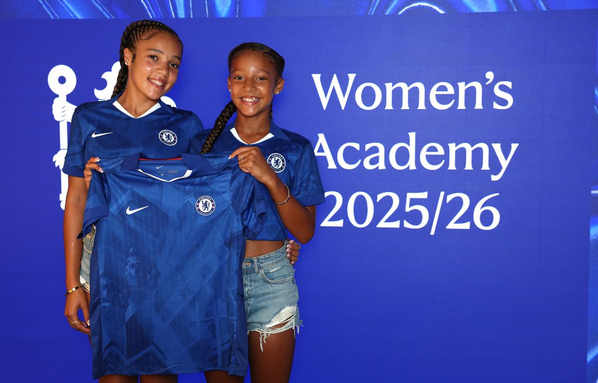 Two young players in Chelsea kit holding up a shirt in front of a Chelsea Women’s Academy screen
