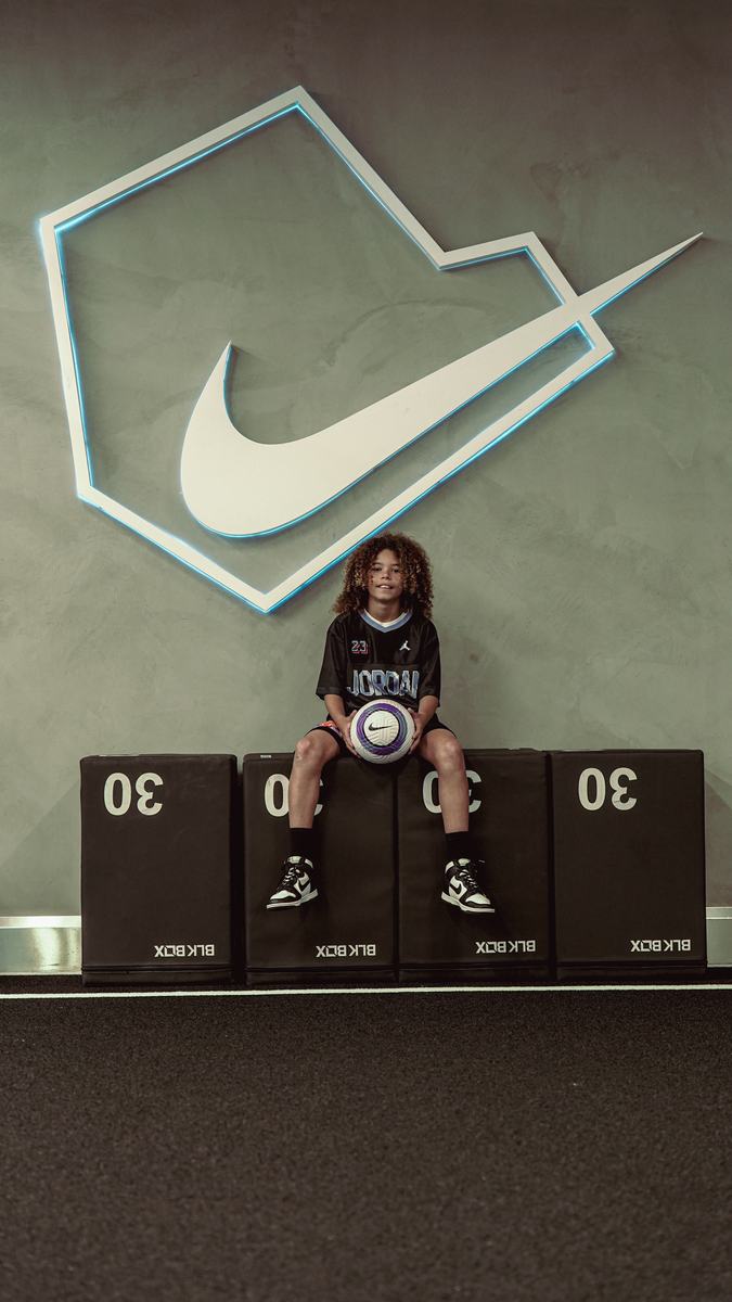 Young player sitting on plyo boxes holding a football beneath a large Nike logo