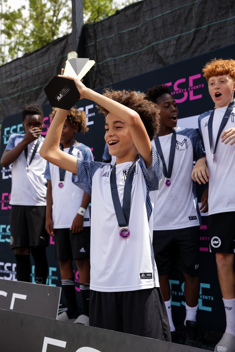 Young player in a Monster Mentality Sport shirt and medal holding a trophy above his head on a podium, teammates celebrating behind him
