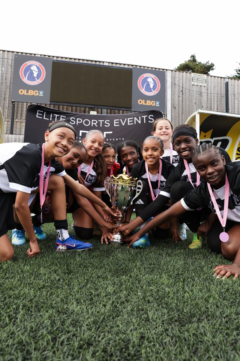 Girls in Monster Mentality Sport kit and pink medals crouching around a silver trophy